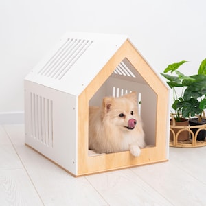 May include: A small, light-colored dog with fluffy fur rests inside a house-shaped pet bed. The structure features white side panels with vertical slats and a natural light wood frame around the front opening, creating a cozy enclosure.