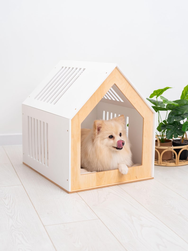May include: A small, light-colored dog with fluffy fur rests inside a house-shaped pet bed. The structure features white side panels with vertical slats and a natural light wood frame around the front opening, creating a cozy enclosure.