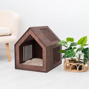 May include: A dark brown wooden dog house with a house-shaped design. The dog house has a slatted roof and side panels, and a beige cushion inside. A potted plant in a woven basket sits nearby.