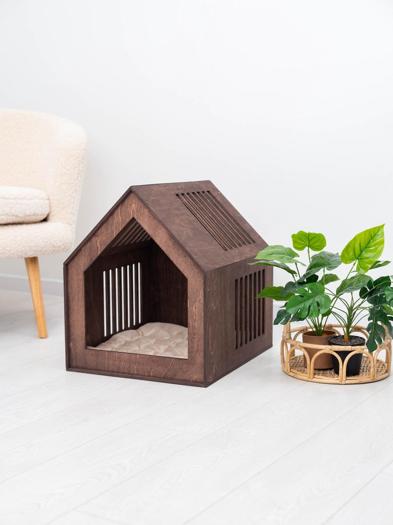 May include: A dark brown wooden dog house with a house-shaped design. The dog house has a slatted roof and side panels, and a beige cushion inside. A potted plant in a woven basket sits nearby.