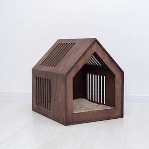 May include: A wooden pet house in a house shape, stained a dark brown color. The house has a slatted roof and side panels, with an open front and a beige cushion inside. The pet house is designed for small dogs or cats.