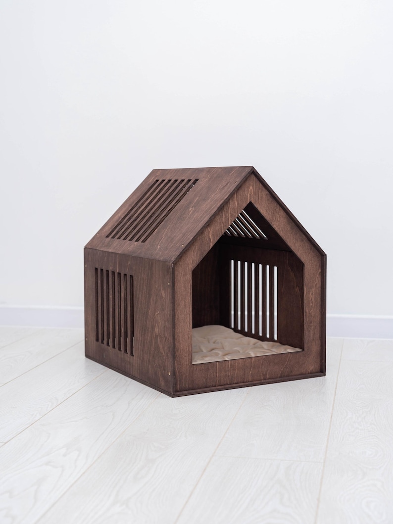May include: A wooden pet house in a house shape, stained a dark brown color. The house has a slatted roof and side panels, with an open front and a beige cushion inside. The pet house is designed for small dogs or cats.