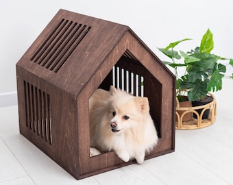 May include: A small, white dog sits inside a wooden dog house with a slatted front. The house is painted a dark brown and has a peaked roof. The dog is looking to the right of the image.