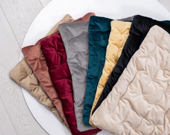 Puede incluir: Una colección de cojines de terciopelo acolchados en varios colores, incluyendo beige, rosa, burdeos, gris, verde azulado, amarillo, negro y crema. Los cojines están dispuestos en forma de abanico sobre una superficie blanca, mostrando su textura suave y ricos tonos.