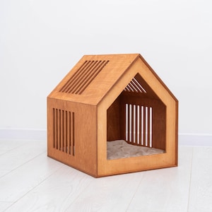 May include: A wooden pet house with a gabled roof and slatted walls. The structure features a large triangular front opening and a light-colored, quilted fabric cushion placed on the floor inside the enclosure for comfort.