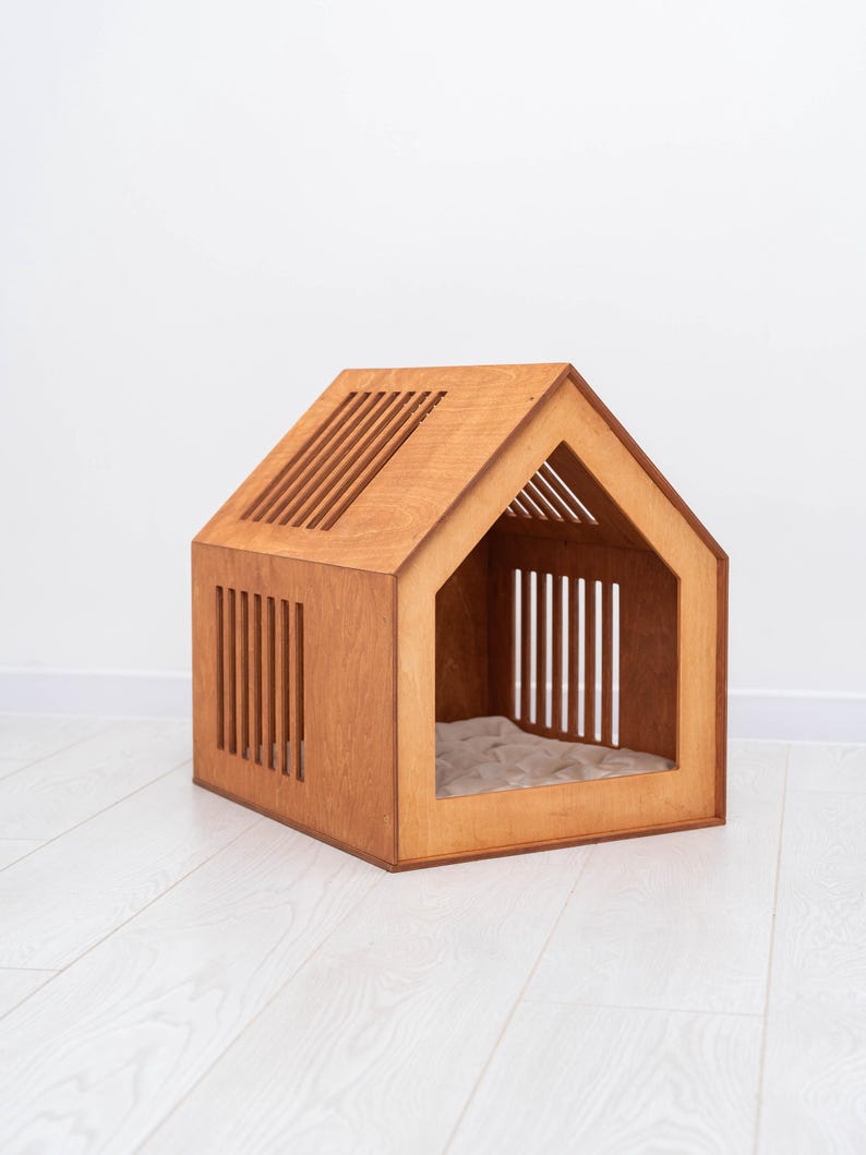 May include: A wooden pet house with a gabled roof and slatted walls. The structure features a large triangular front opening and a light-colored, quilted fabric cushion placed on the floor inside the enclosure for comfort.