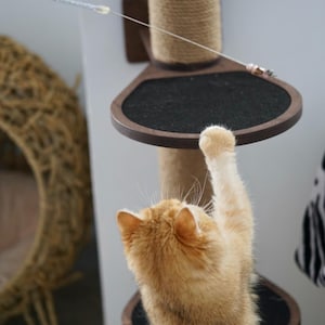 May include: A ginger cat reaches up to a toy on a string hanging from a cat tree. The cat tree is made of wood and sisal rope. The cat is standing on a platform with a black surface.