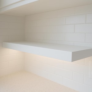 May include: A white floating shelf is mounted on a white tiled wall. The shelf is rectangular and has a clean, modern design. A light strip is installed under the shelf, illuminating the countertop below. An electrical outlet is visible on the right.