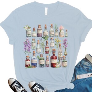 May include: Light blue t-shirt with a vintage illustration of various glass bottles filled with colorful liquids and flowers. The bottles have labels with handwritten text.