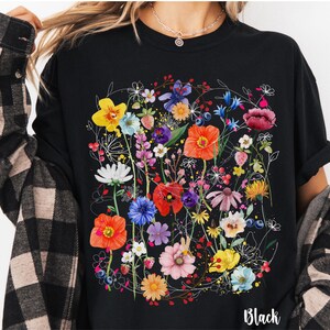 May include: Black t-shirt with a colorful floral print. The print features a variety of flowers, including poppies, daisies, and wildflowers. The shirt has a crew neck and short sleeves. The text "Black" is printed on the bottom of the shirt.