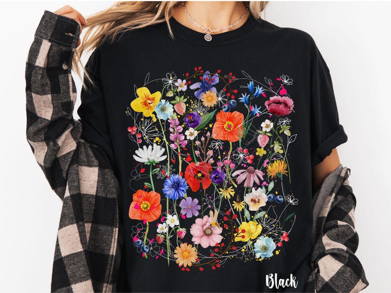 May include: Black t-shirt with a colorful floral print. The print features a variety of flowers, including poppies, daisies, and wildflowers. The shirt has a crew neck and short sleeves. The text "Black" is printed on the bottom of the shirt.