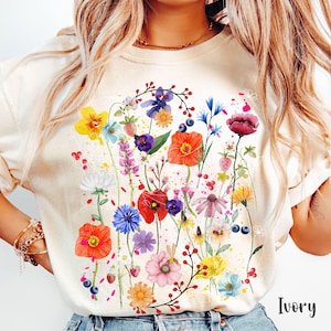 May include: A cream colored t-shirt with a colorful floral print. The print features a variety of flowers, including red, pink, yellow, blue, and purple flowers. The shirt has a crew neck and short sleeves. The text "Ivory" is visible in the bottom right corner of the image.