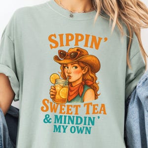 May include: A light blue t-shirt with a graphic of a woman wearing a cowboy hat and sunglasses, drinking sweet tea. The text on the shirt reads "Sippin' Sweet Tea & Mindin' My Own".