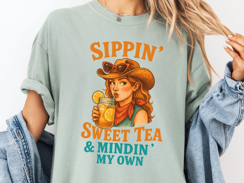 May include: A light blue t-shirt with a graphic of a woman wearing a cowboy hat and sunglasses, drinking sweet tea. The text on the shirt reads "Sippin' Sweet Tea & Mindin' My Own".