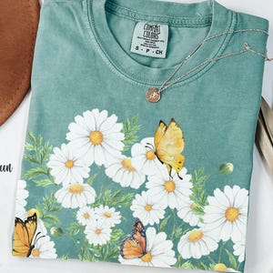 以下が含まれることがあります： デイジーと蝶のグラフィックが特徴の、ライトグリーンのComfort Colors Tシャツ。クルーネックと半袖です。小さなペンダントが付いたシルバーのネックレスがシャツの上にかけられています。