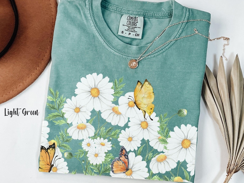 以下が含まれることがあります： デイジーと蝶のグラフィックが特徴の、ライトグリーンのComfort Colors Tシャツ。クルーネックと半袖です。小さなペンダントが付いたシルバーのネックレスがシャツの上にかけられています。