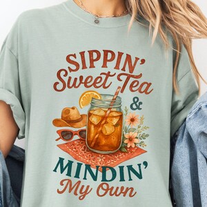 Może przedstawiać: Jasno błękitny t-shirt z nadrukiem graficznym przedstawiającym szklankę lodowatej herbaty, kowbojski kapelusz, okulary przeciwsłoneczne i kwiaty. Tekst na koszulce brzmi "Sippin' Sweet Tea & Mindin' My Own".