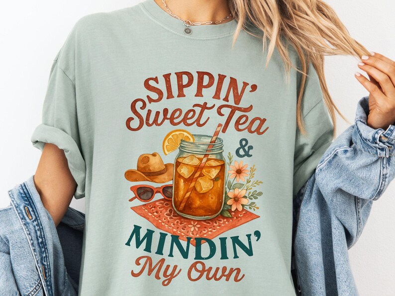 Może przedstawiać: Jasno błękitny t-shirt z nadrukiem graficznym przedstawiającym szklankę lodowatej herbaty, kowbojski kapelusz, okulary przeciwsłoneczne i kwiaty. Tekst na koszulce brzmi "Sippin' Sweet Tea & Mindin' My Own".