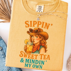 May include: A yellow t-shirt with a cartoon illustration of a woman wearing a cowboy hat and sunglasses, sipping sweet tea. The text "Sippin' Sweet Tea & Mindin' My Own" is printed on the shirt.