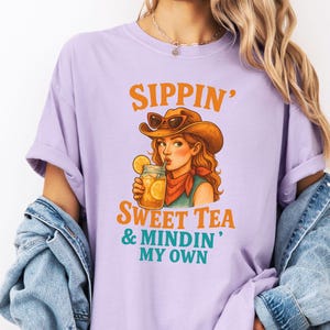 May include: A purple t-shirt with the text "Sippin' Sweet Tea & Mindin' My Own" and a cartoon illustration of a woman wearing a cowboy hat and sunglasses, drinking a glass of iced tea.