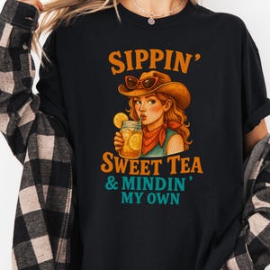 May include: Black t-shirt with a graphic of a woman wearing a cowboy hat and drinking sweet tea. The text "Sippin' Sweet Tea & Mindin' My Own" is printed on the shirt.