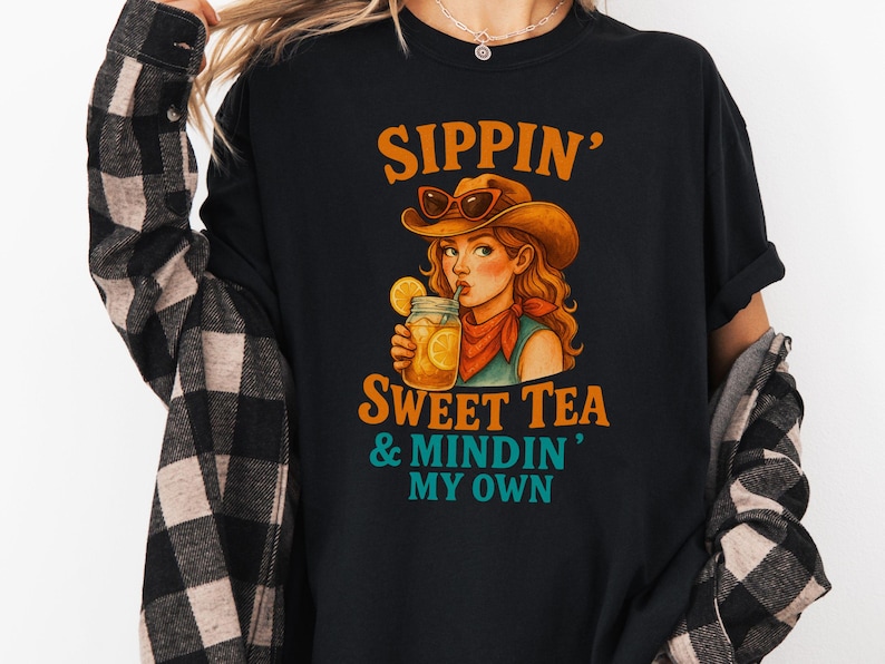 May include: Black t-shirt with a graphic of a woman wearing a cowboy hat and drinking sweet tea. The text "Sippin' Sweet Tea & Mindin' My Own" is printed on the shirt.
