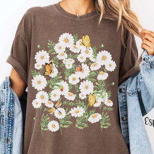 以下が含まれることがあります： エスプレッソ色のTシャツ。白いデイジー、緑の葉、蝶のフローラルデザイン。蝶は黄色とオレンジ色です。シャツはゆったりとしたフィット感で、袖はロールアップされています。