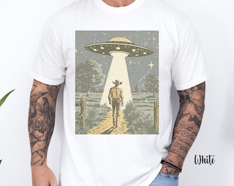 May include: A white t-shirt with a vintage-style illustration of a cowboy walking towards a flying saucer in a field. The illustration is in shades of brown, green, and blue.