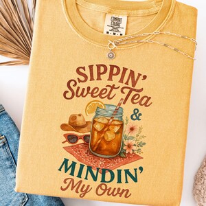 Może przedstawiać: Ż&oacute;łta koszulka z napisem "Sippin' Sweet Tea & Mindin' My Own" w brązowych literach. Obok tekstu znajduje się szklanką lodowatej herbaty z plasterkiem cytryny i słomką.