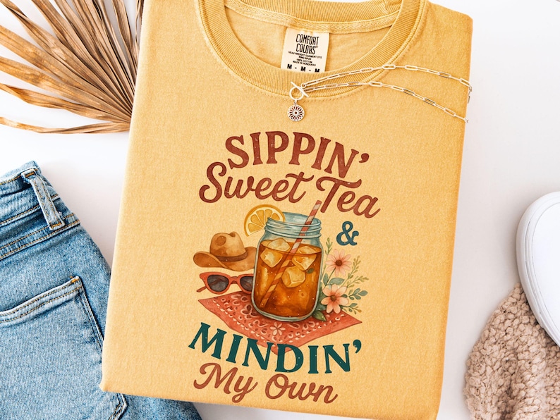 Może przedstawiać: Ż&oacute;łta koszulka z napisem "Sippin' Sweet Tea & Mindin' My Own" w brązowych literach. Obok tekstu znajduje się szklanką lodowatej herbaty z plasterkiem cytryny i słomką.