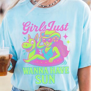 May include: Light blue t-shirt with a graphic of a woman in a swimsuit and cowboy hat floating on an inner tube with a cartoon horse. The text "Girls Just Wanna Have Sun" is in pink and green.
