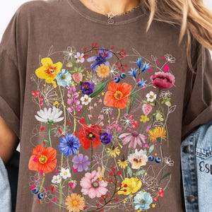 May include: A brown t-shirt with a colorful floral print. The print features a variety of flowers, including red poppies, blue cornflowers, and yellow daisies. The flowers are arranged in a loose, organic pattern.