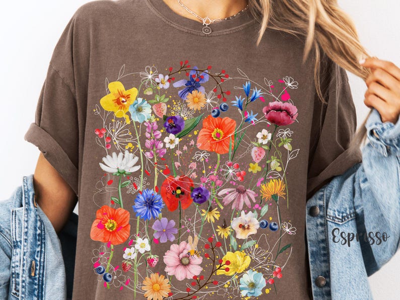 May include: A brown t-shirt with a colorful floral print. The print features a variety of flowers, including red poppies, blue cornflowers, and yellow daisies. The flowers are arranged in a loose, organic pattern.