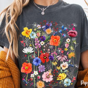 May include: A gray t-shirt with a floral print featuring a variety of colorful flowers, including red, yellow, pink, blue, and purple. The flowers are arranged in a cluster and are surrounded by green leaves.