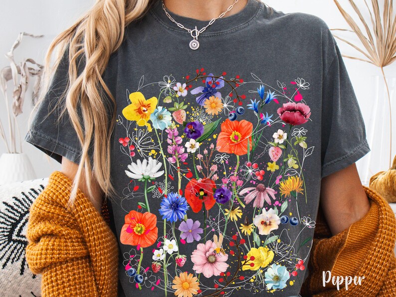 May include: A gray t-shirt with a floral print featuring a variety of colorful flowers, including red, yellow, pink, blue, and purple. The flowers are arranged in a cluster and are surrounded by green leaves.
