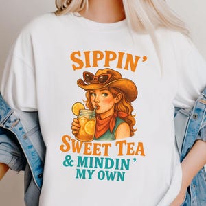 May include: White t-shirt with a cartoon illustration of a woman wearing a cowboy hat and drinking sweet tea. The text "Sippin' Sweet Tea & Mindin' My Own" is printed on the shirt.