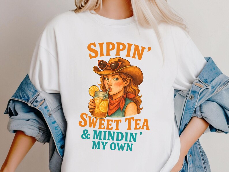 May include: White t-shirt with a cartoon illustration of a woman wearing a cowboy hat and drinking sweet tea. The text "Sippin' Sweet Tea & Mindin' My Own" is printed on the shirt.