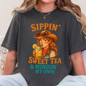 May include: A dark gray t-shirt with a graphic of a woman wearing a cowboy hat and drinking sweet tea. The text on the shirt reads "Sippin' Sweet Tea & Mindin' My Own."