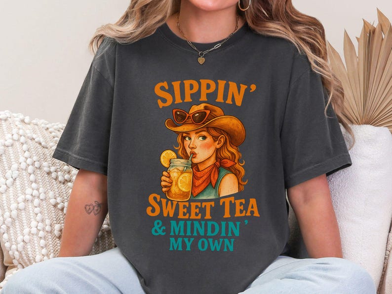 May include: A dark gray t-shirt with a graphic of a woman wearing a cowboy hat and drinking sweet tea. The text on the shirt reads "Sippin' Sweet Tea & Mindin' My Own."