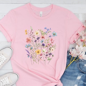 May include: A pink t-shirt with a floral print featuring a variety of colorful flowers, including yellow, blue, pink, and purple.