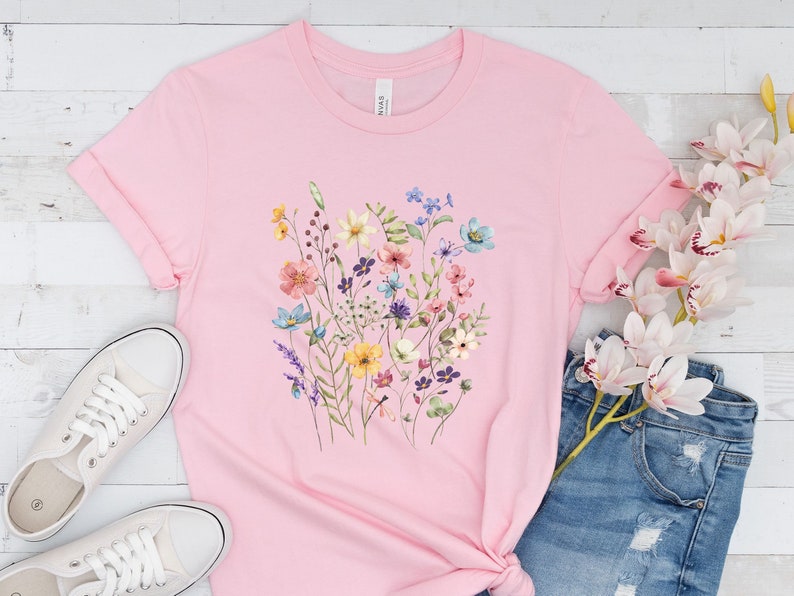 May include: A pink t-shirt with a floral print featuring a variety of colorful flowers, including yellow, blue, pink, and purple.