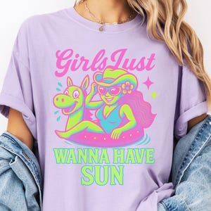 May include: Lilac t-shirt with a graphic of a woman in a swimsuit and sunglasses floating on a pink inner tube with a horse-shaped float. Text reads "Girls Just Wanna Have Sun" in pink and green.