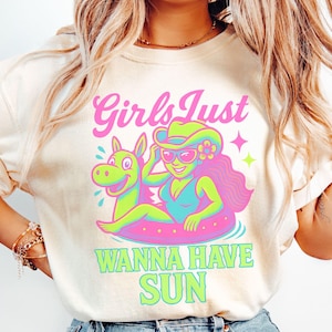 May include: Cream-colored t-shirt with a graphic featuring a cartoon woman in a swimsuit and cowboy hat, floating on a pink inner tube with a green horse. The text reads "Girls Just Wanna Have Sun."