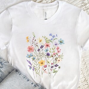May include: White t-shirt with a watercolor floral print featuring pink, blue, yellow, and purple flowers.