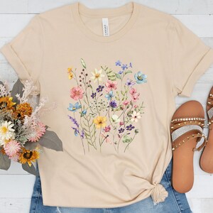 May include: A beige t-shirt with a floral print featuring a variety of colorful flowers, including pink, blue, yellow, and purple.