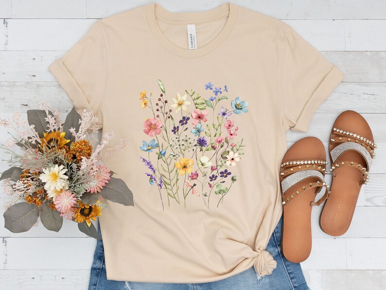 May include: A beige t-shirt with a floral print featuring a variety of colorful flowers, including pink, blue, yellow, and purple.