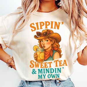 May include: A white t-shirt with a graphic of a woman wearing a cowboy hat and sunglasses, holding a glass of iced tea. The text on the shirt reads "Sippin' Sweet Tea & Mindin' My Own".