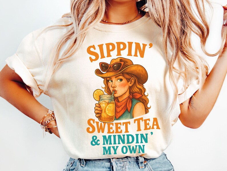 May include: A white t-shirt with a graphic of a woman wearing a cowboy hat and sunglasses, holding a glass of iced tea. The text on the shirt reads "Sippin' Sweet Tea & Mindin' My Own".