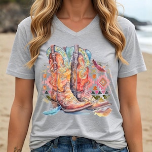 May include: A gray V-neck t-shirt featuring a watercolor illustration of two cowboy boots. The boots are in shades of orange, yellow, and brown, with blue and green accents. A colorful geometric pattern and feathers are also present.