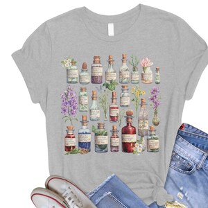 May include: A gray t-shirt with a design of various glass bottles filled with colorful liquids and herbs. The bottles are surrounded by flowers and leaves.